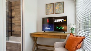 A modern bathroom with a glass shower enclosure, a wooden vanity with a flat-screen TV displaying streaming service logos, and a cozy seating area with a cushion and a vase of flowers. - East Side #212 xxxxxxx (Austin)