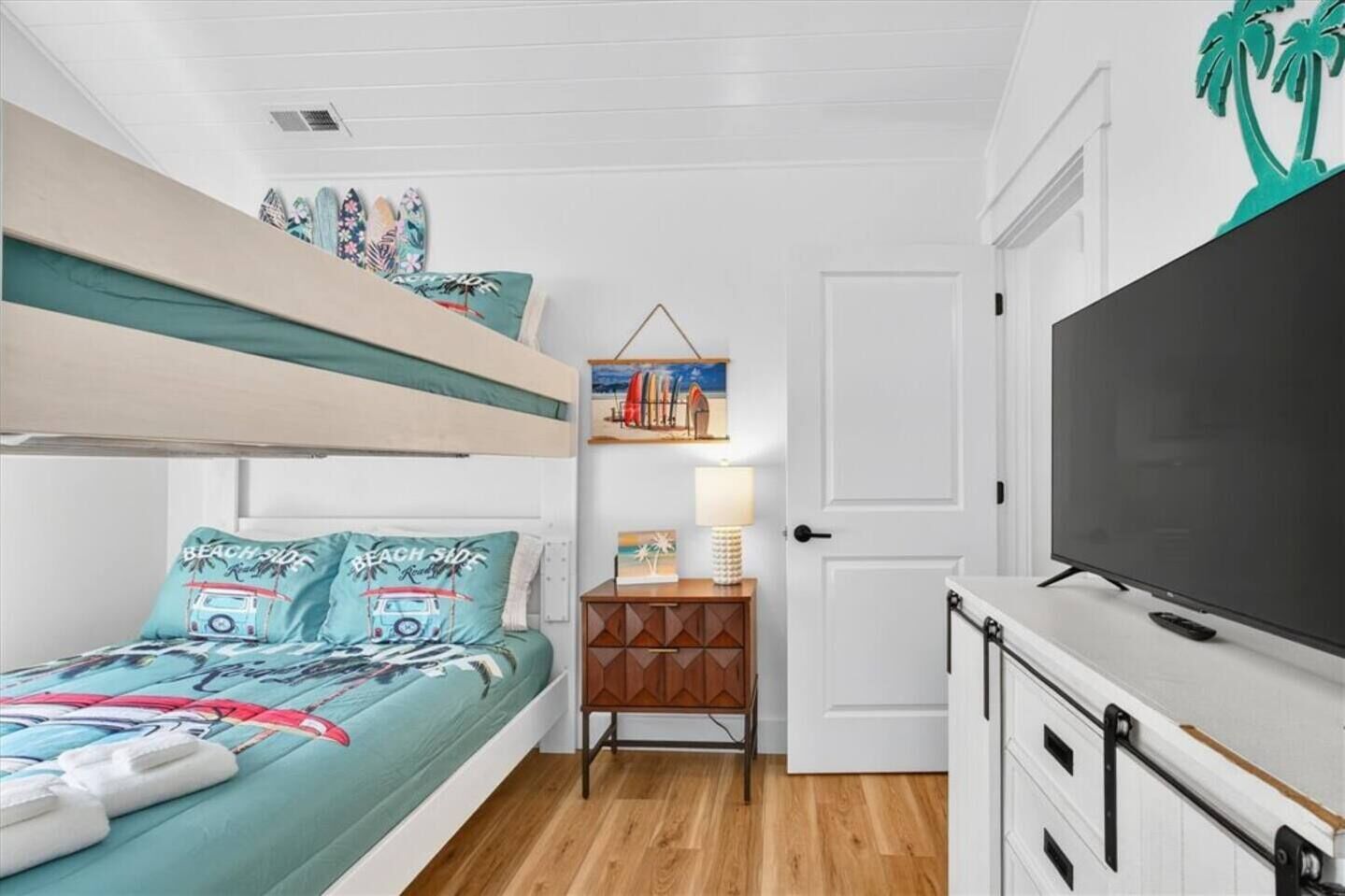 A compact bedroom with a bunk bed, a bedside table with a lamp, a TV, and a door with a palm tree decoration.
