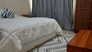 Un dormitorio con una cama, una mesita con una botella, una puerta de madera y un tapete estampado.