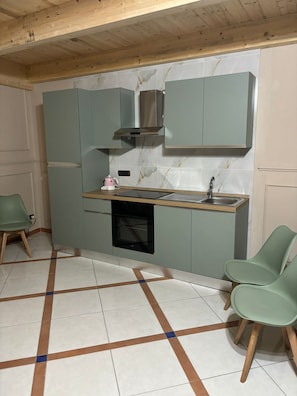 A compact kitchen with a built-in oven, a sink, and a stainless steel range hood. There are two green chairs and a small table.