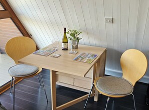 A small dining table with two chairs, a bottle of wine, a vase with flowers, and a magazine on the table.