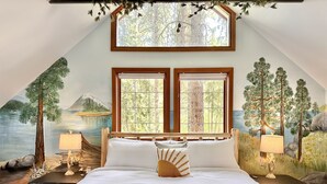 A bedroom with a large bed, two windows, a mural of a forest scene, and a sloped ceiling. - Blue Pine Forest Lodge 3 Bedroom Cabin (Leavenworth)