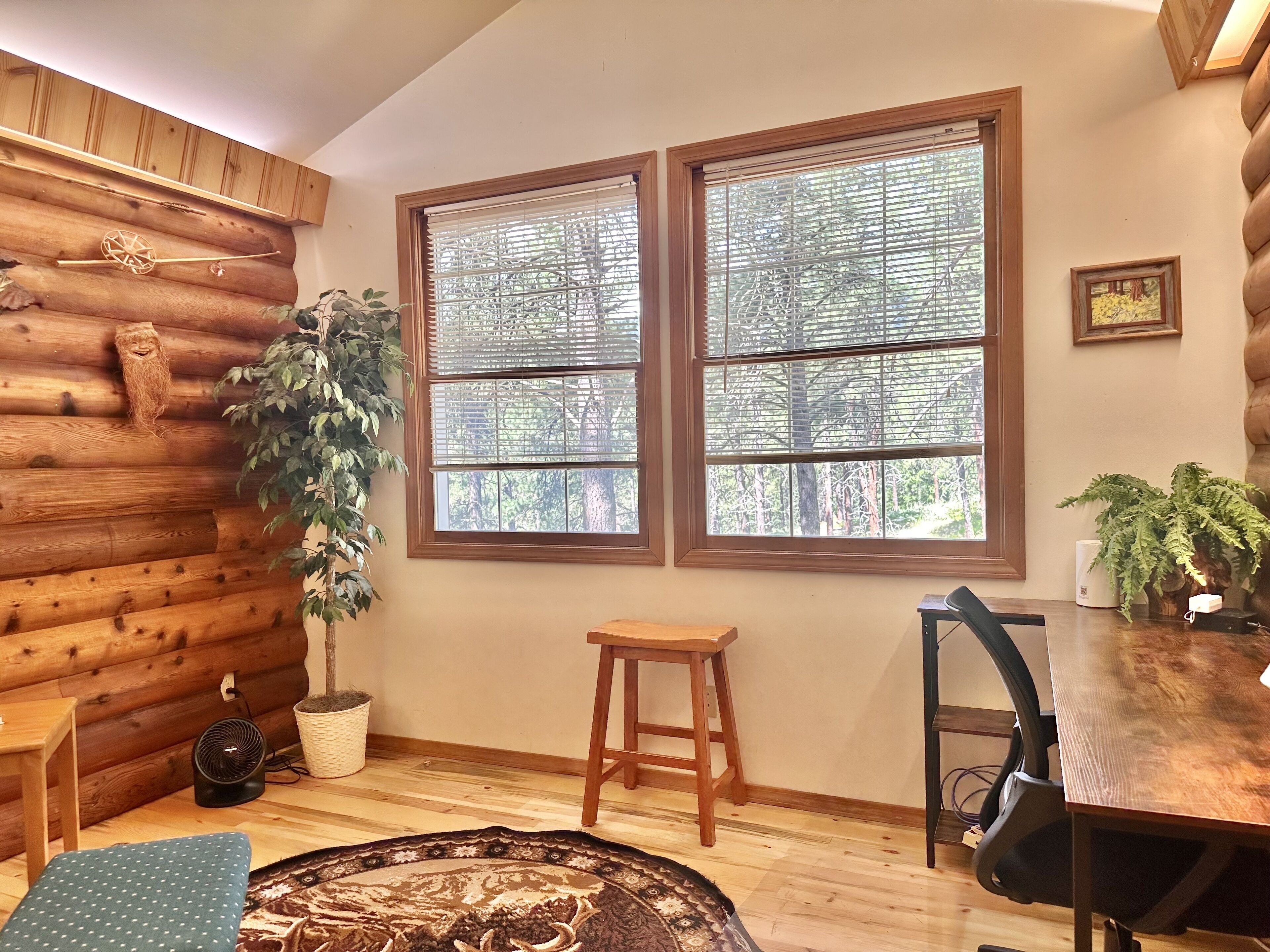 A cozy cabin interior with a log wall, two large windows with blinds, a wooden table, a chair, a small stool, and a potted plant.