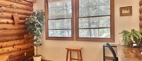 A cozy cabin interior with a log wall, two large windows with blinds, a wooden table, a chair, a small stool, and a potted plant.