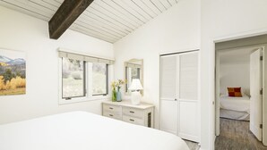Ein Schlafzimmer mit einem großen Bett, einem Schminktisch mit Spiegel, einem Fenster mit Blick auf Bäume und einer Tür zu einem weiteren Raum.