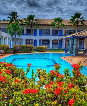 Una piscina rodeada de palmeras y flores rojas, con un edificio de ventanas azules y techo de tejas al fondo.