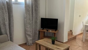 A living room with a sofa, a TV on a wooden stand, a small table with a plant, patterned curtains, and an air conditioning unit. - T2 apartment in condominium (Villa) with pool access (Fréjus)