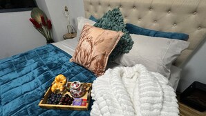 A neatly made bed with a blue quilt, a white fluffy blanket, and a tray with croissants, tea, and fruit.