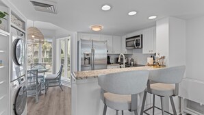 Een moderne keuken met witte kasten, roestvrijstalen apparatuur, een centraal eiland met barkrukken en uitzicht op het buiten.