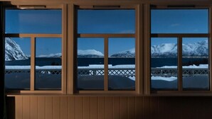 A room with a large window overlooking a snowy landscape with mountains and a body of water. - Lyngen Aurora House (Lyngseidet)