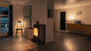 A cozy living room with a wood-burning stove, a chair, and a dining area with a cabinet and a clock on the wall. - Lyngen Aurora House (Lyngseidet)