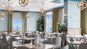 A hotel dining area with multiple tables and chairs, large windows with curtains, and hanging light fixtures.