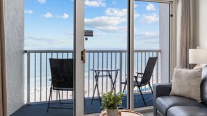 A balcony with a view of the beach, a sofa, two chairs, and a small table with a plant.