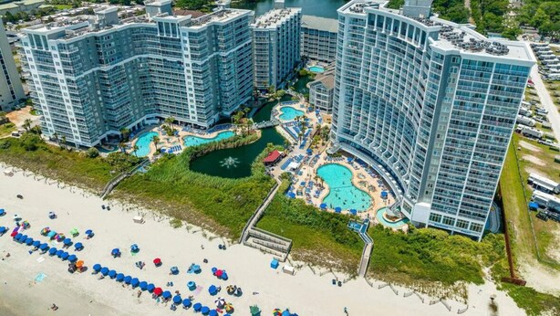 A high-rise hotel with multiple swimming pools, a beach area with umbrellas, and a view of the surrounding greenery.