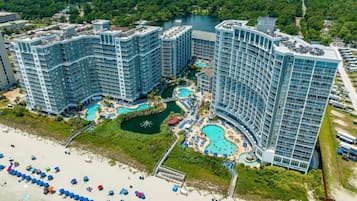 Un hotel de gran altura con múltiples piscinas, una zona de playa con sombrillas y vistas al entorno verde.