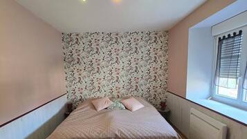 Un dormitorio con cama, papel tapiz floral, un radiador y una ventana con persianas.