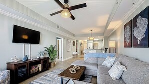 A spacious living room with a large gray sofa, a wooden coffee table, and a ceiling fan. A flat-screen TV is mounted on the wall, and there are decorative items on a console table. - Disney Coastal Condo (Orlando)