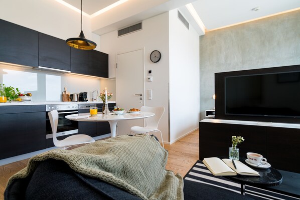 A modern kitchen with dark cabinets, a dining area with a round table, and a living space with a TV and a coffee table.
