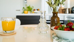 Una escena de desayuno con un vaso de jugo de naranja, un florero decorativo con flores y un bol de fresas sobre una mesa.