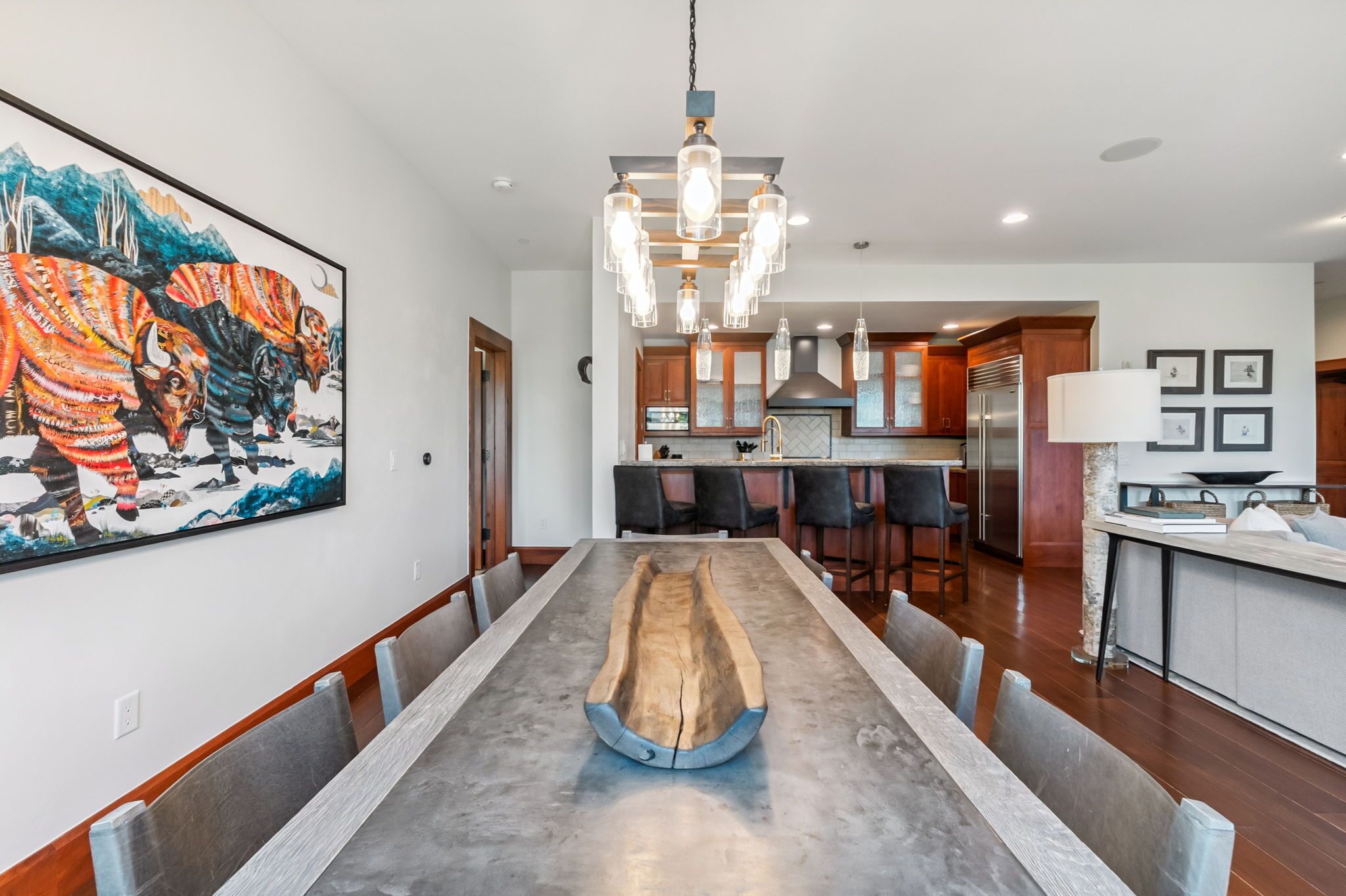 A modern dining room with a long table, a kitchen in the background, and a large painting on the wall.