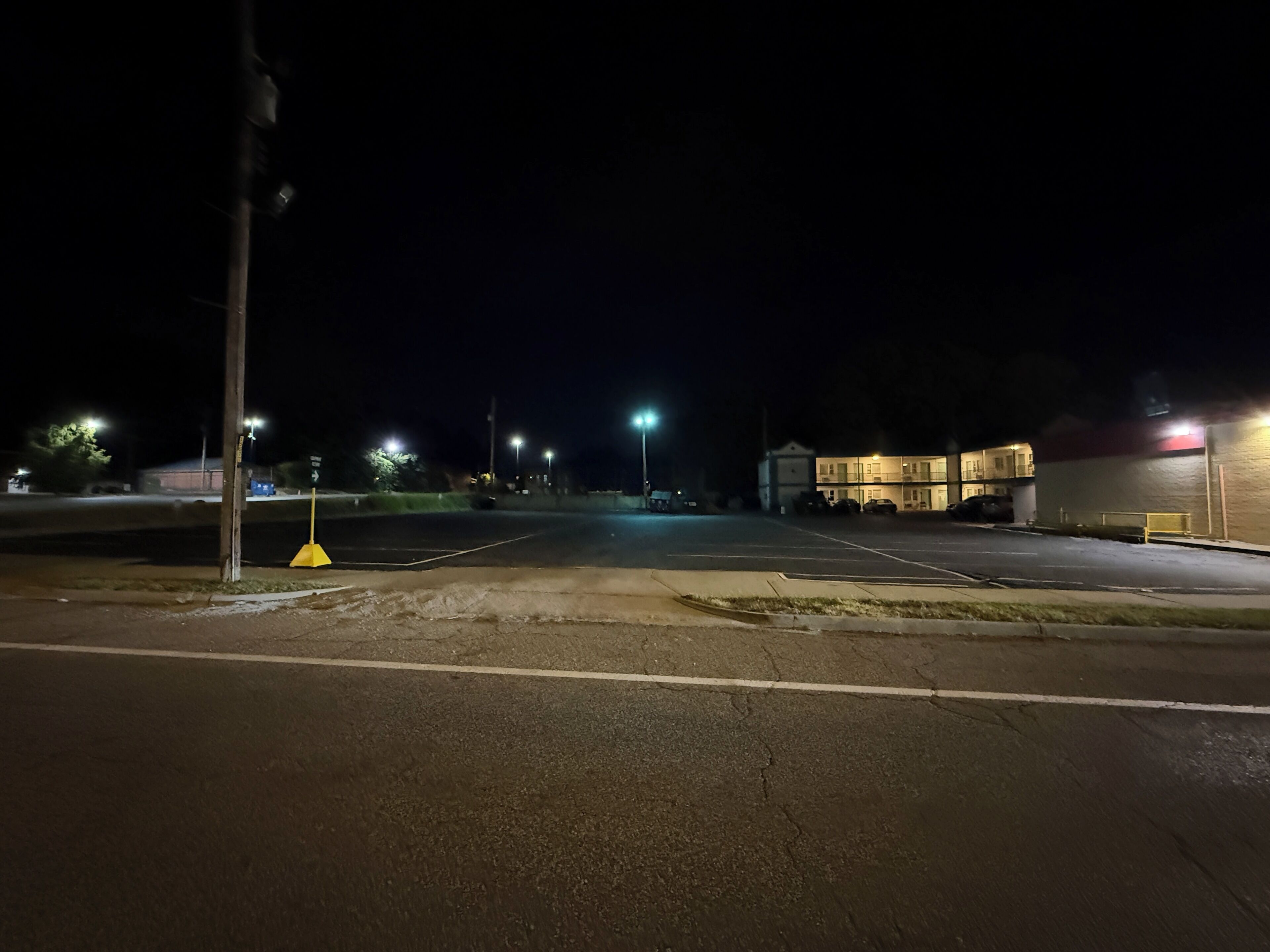 Un estacionamiento nocturno con algunos coches, farolas y un edificio al fondo.