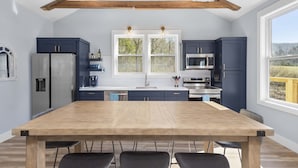 A modern kitchen with dark blue cabinets, a wooden dining table, and a large window providing a view of the outdoors. - Creekside Retreat • Fire Pit | Patio | Arcade (Old Fort)