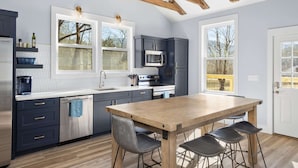 A modern kitchen with dark cabinets, a wooden dining table, and a large window providing a view of trees. - Creekside Retreat • Fire Pit | Patio | Arcade (Old Fort)