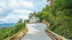A house with a paved driveway leading to a wooden gate, surrounded by lush greenery and a mountainous backdrop.