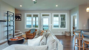 A spacious living room with a large sectional sofa, a coffee table, and a dining area with chairs. There are large windows with a view of the ocean, a staircase, and a bookshelf.