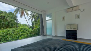 A modern room with a large window overlooking lush greenery, a black rug, and a wall-mounted TV. - Colonial-style bungalow with private pool, mountain views  and tranquil vibes. (Kandy)
