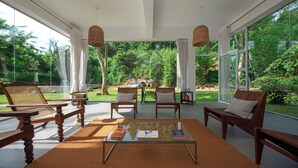 A spacious living area with a glass coffee table, wooden chairs, and a view of a lush garden through large windows. - Colonial-style bungalow with private pool, mountain views  and tranquil vibes. (Kandy)