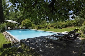 Una piscina rectangular con borde de piedra, rodeada por un patio de piedra. Se observan sillones de descanso y un paraguas.