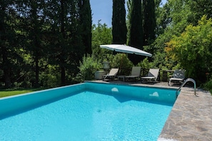 A rectangular swimming pool with clear blue water, surrounded by stone steps and a lounge area with chairs and an umbrella.