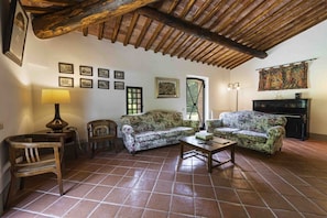 Una sala de estar con techo de madera, suelo de baldosas, sofás con estampado floral, una mesa de madera y una chimenea.