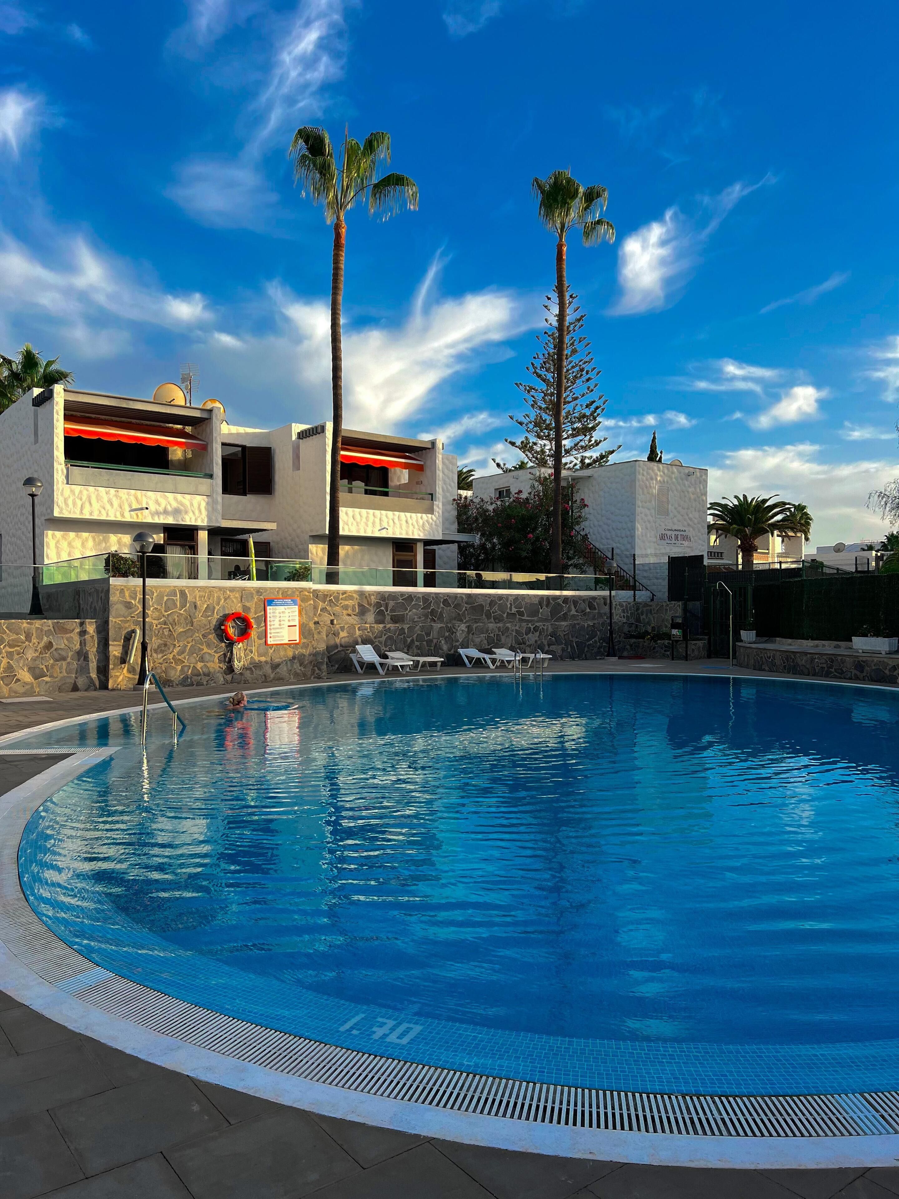 A swimming pool with lounge chairs, a stone wall, and modern buildings with balconies.