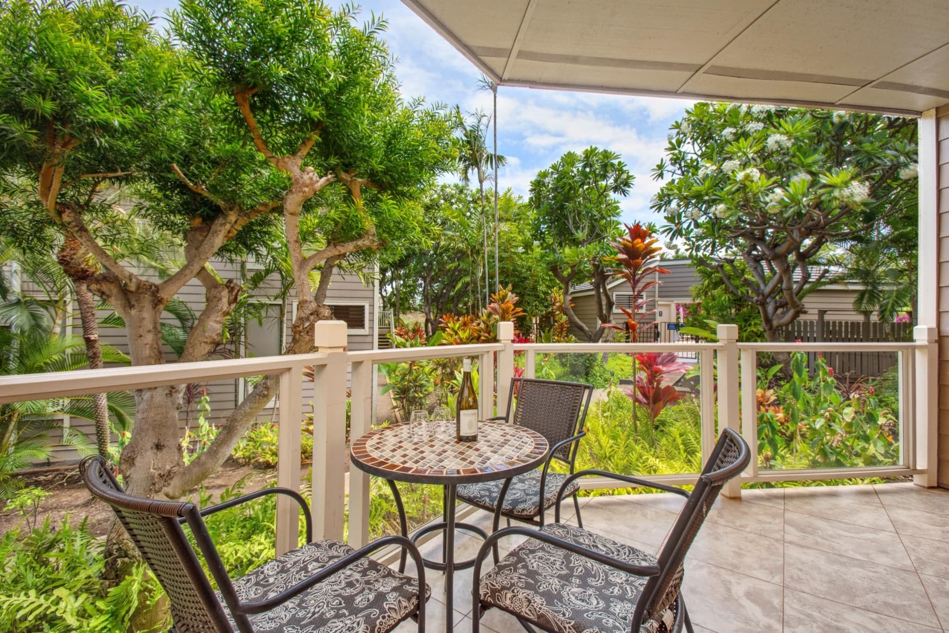 A covered outdoor patio with a round table and four chairs, surrounded by lush greenery and trees.
