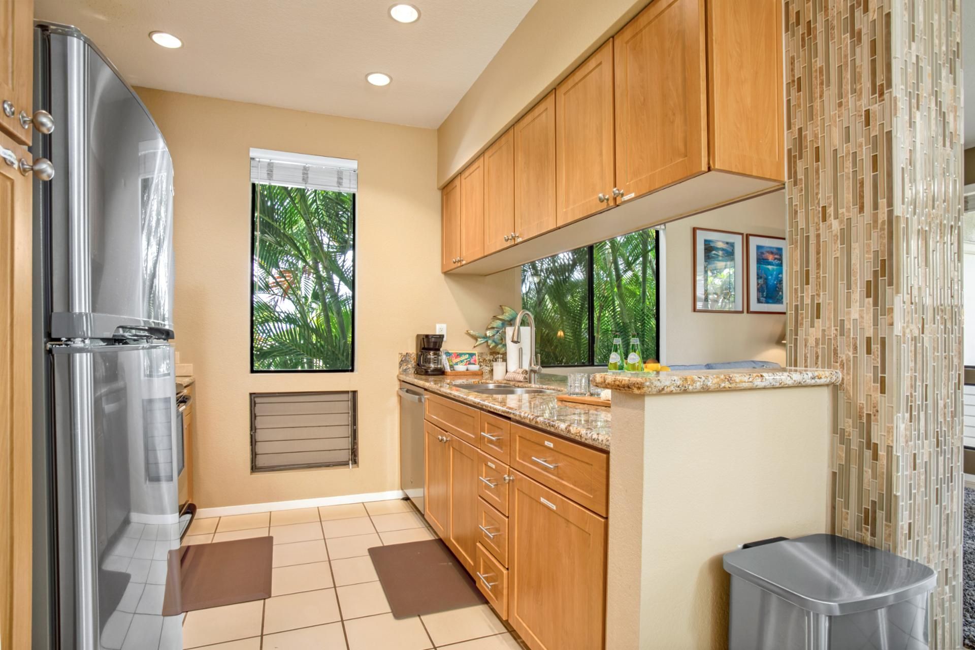 A modern kitchen with wooden cabinets, a stainless steel refrigerator, a window with a view of trees, and a tiled floor.
