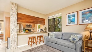 A modern living room with a grey sofa, two wooden stools, a patterned rug, and a kitchen with wooden cabinets and stainless steel appliances.