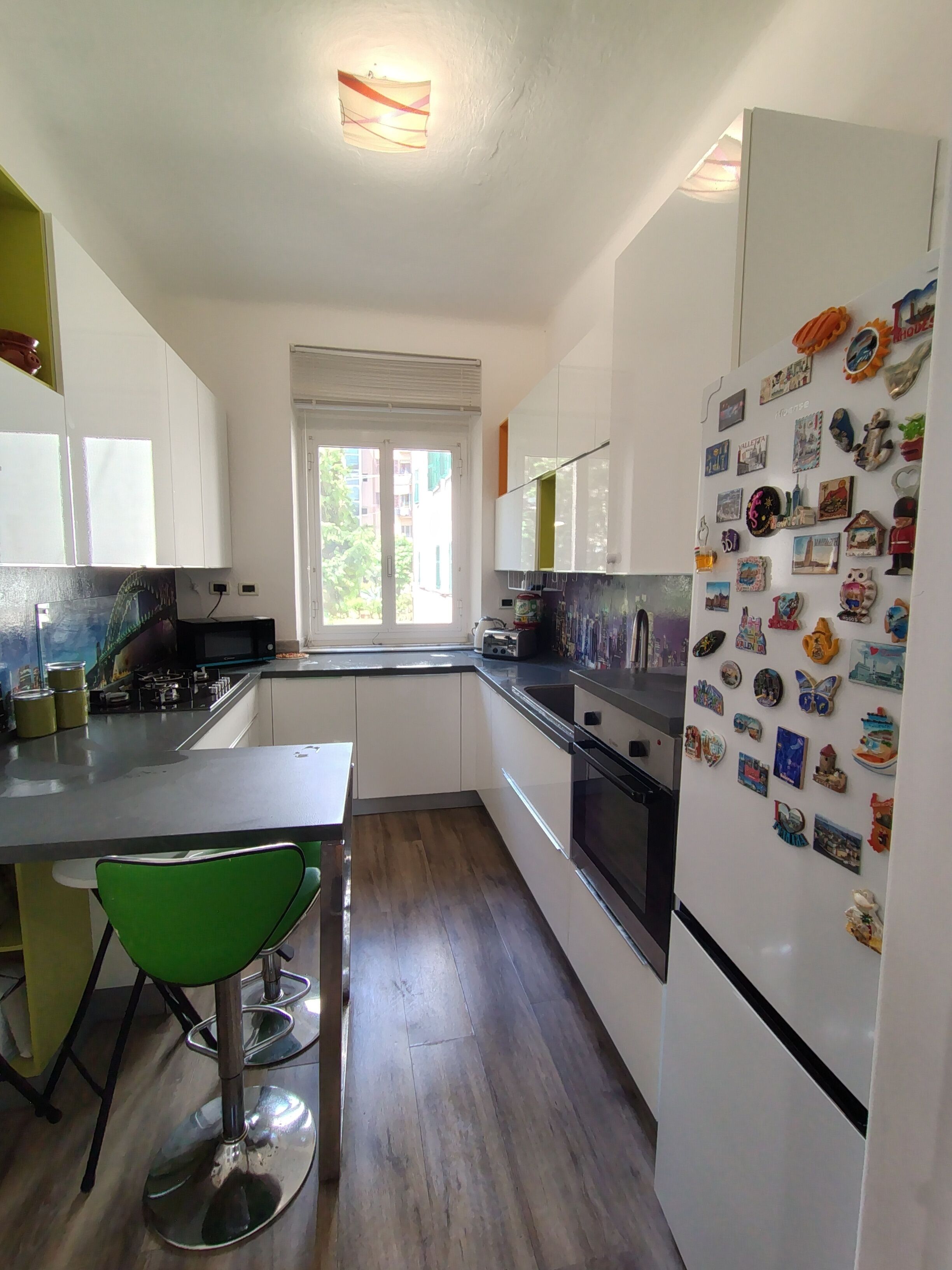 A compact kitchen with white cabinets, a refrigerator adorned with magnets, a window with a view of buildings, and a green chair.