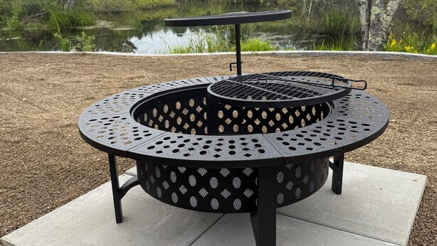 Ein runder Grill für den Außenbereich mit einem Metallrost, auf einer Betonplatte, umgeben von Kies.