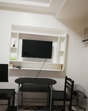 A room with a TV mounted on a white wall, a dining table with chairs, and a fan on the floor. - Grand Riviera Condotel Near St Lukes Medical Center (Manila)