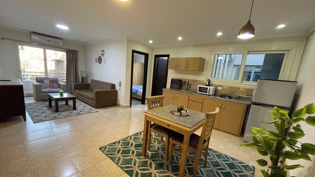 A modern apartment with a living room, dining area, and kitchen. It features a large window, a dining table with chairs, a kitchen with cabinets and appliances, and a potted plant. - Royal Residence Cairo (Giza)