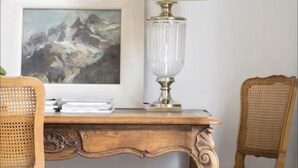 A wooden desk with a glass lamp, a framed painting of mountains, and wicker chairs.