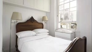 A neatly made bed with white linens, two bedside lamps, and a window with a view of buildings.
