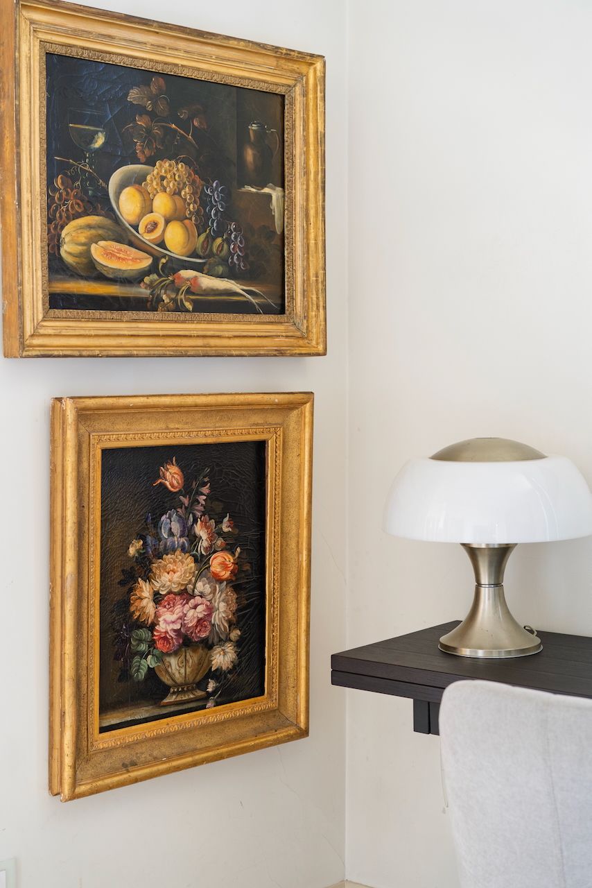 Two framed still life paintings on a wall, a lamp on a table beside them.