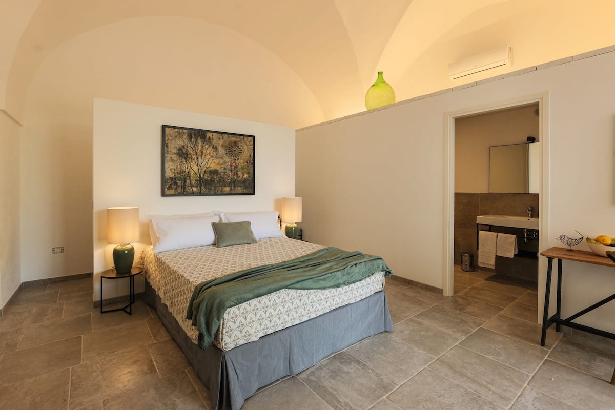 Quarto com cama, abajures, um quadro, um vaso e um banheiro visível por uma porta aberta.
