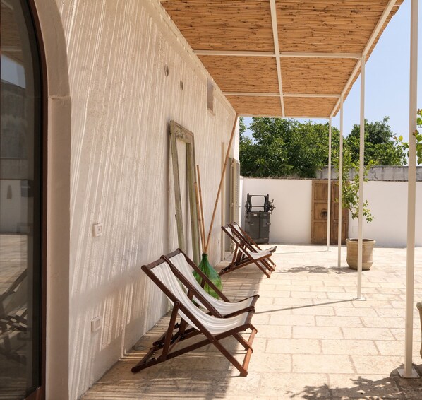 A covered outdoor seating area with wooden chairs and a table, a wooden ceiling, and a white wall with a textured pattern.