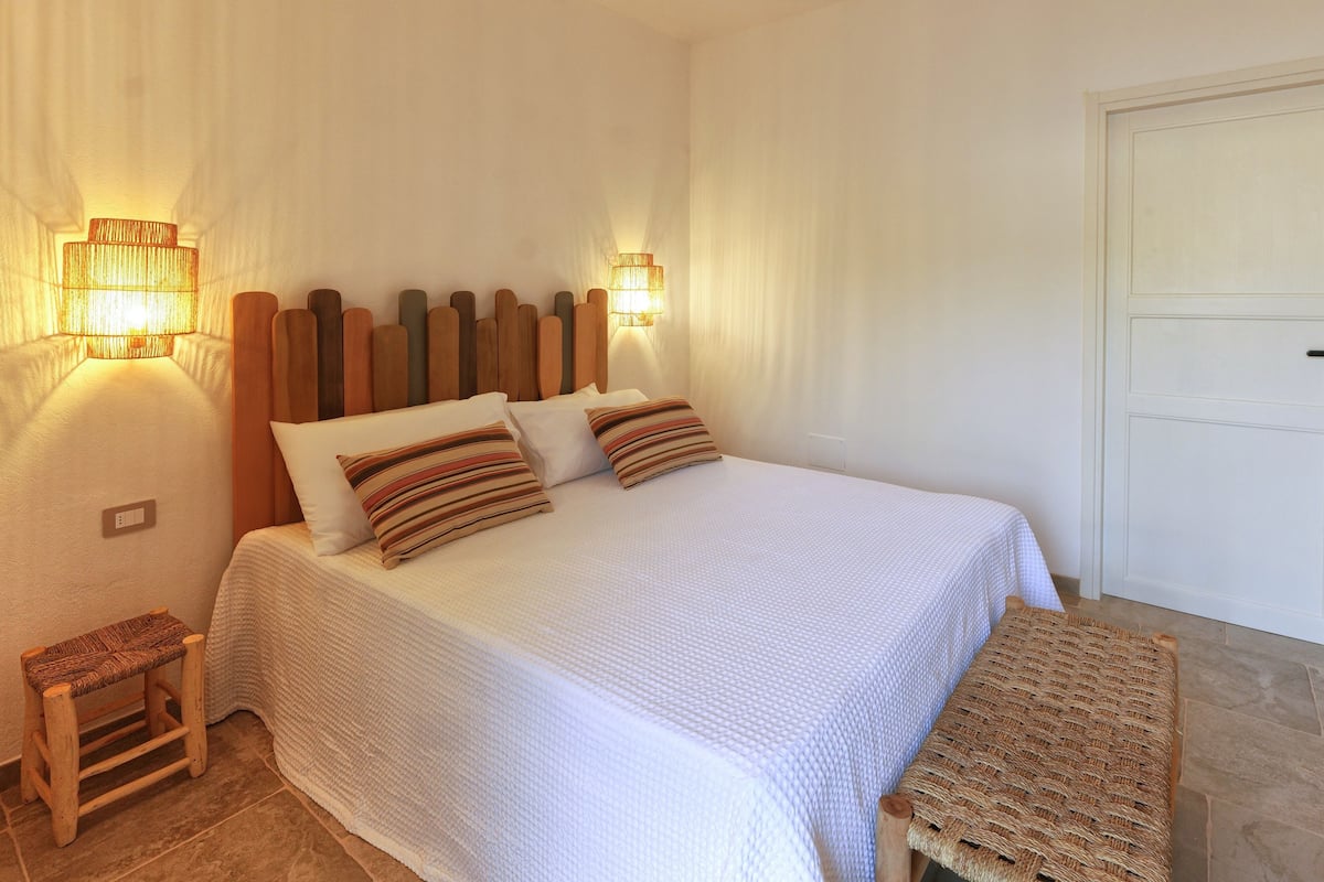 Cama bem arrumada com colcha branca e almofadas listradas, cabeceira de madeira e duas luminárias fixadas na parede.