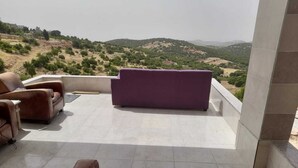 Una terrassa amb un sofà morat i dues cadires de pell marró, amb vista a un paisatge amb turons.