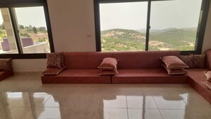 Àmplia zona d'estar amb un sofà vermell gran, una taula petita i vista a un paisatge amb turons a través de grans finestres.
